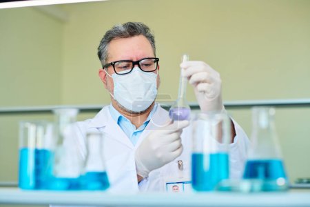 Foto de Químico o farmacólogo masculino maduro serio en anteojos, bata de laboratorio y máscara protectora que sostiene el vaso de precipitados con sustancia líquida en laboratorio - Imagen libre de derechos