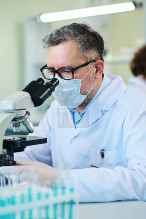 Photo for Mature expert in virology wearing lab coat, eyeglasses, protective mask and gloves studying cells of new virus in microscope in laboratory - Royalty Free Image