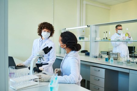 Téléchargez les photos : Deux virologistes féminines interculturelles en blouse de laboratoire et masques protecteurs discutant des caractéristiques du nouveau virus en laboratoire - en image libre de droit