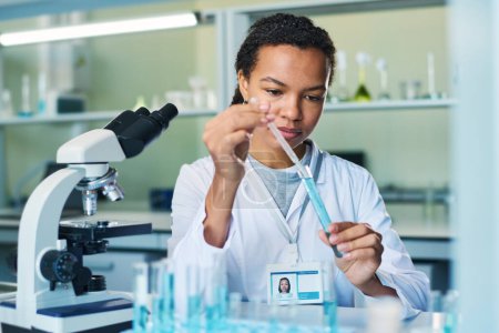 Foto de Farmacóloga joven con gotero que toma muestras de sustancia líquida azul del frasco durante el experimento clínico en laboratorio - Imagen libre de derechos