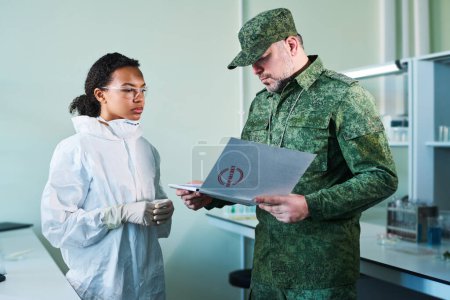 Foto de Oficial maduro en uniforme de camuflaje mirando a través de notas que describen cada etapa de experimento clínico secreto en el laboratorio militar - Imagen libre de derechos