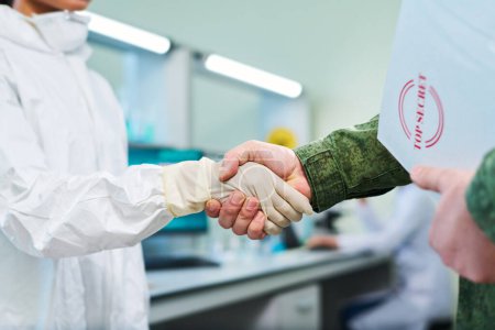 Photo for Focus on handshake of officer and young female clinician wearing white protective gloves and hazmat suit in military laboratory - Royalty Free Image