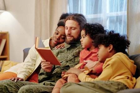 Photo for Young man in military uniform reading book to his adorable sons and affectionate wife surrounding him while sitting on couch - Royalty Free Image