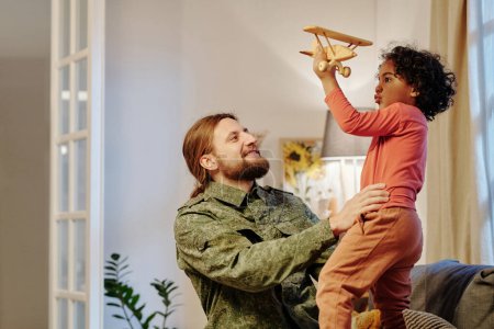 Téléchargez les photos : Joyeux jeune homme en uniforme militaire s'amusant avec son mignon petit fils jouant avec un avion jouet tout en se tenant devant lui à la maison - en image libre de droit