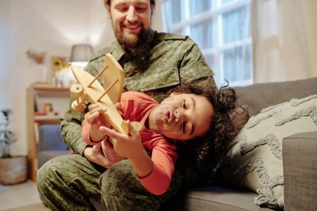 Téléchargez les photos : Adorable petit garçon jouant avec un avion jouet en bois sur les mains de son père tout en s'amusant à loisir sur le canapé dans un environnement familial - en image libre de droit