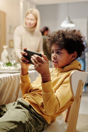 Téléchargez les photos : Adorable garçon afro-américain regardant la vidéo en ligne sur smartphone tout en étant assis sur la chaise contre sa grand-mère table de service pour le dîner - en image libre de droit
