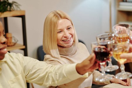 Téléchargez les photos : Joyeux blond mature femme cliquetis avec des verres de vin des invités assis à table festive et profiter d'un dîner en famille en l'honneur de l'événement de la vie - en image libre de droit