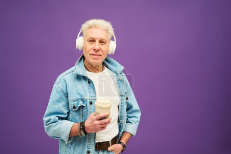 Téléchargez les photos : Homme âgé aux cheveux blancs regardant la caméra tout en tenant une tasse de café et en écoutant de la musique dans un casque sur fond violet - en image libre de droit