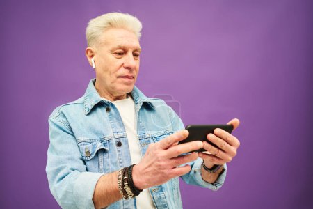 Téléchargez les photos : Homme âgé élégant veste en denim regardant l'écran du smartphone tout en regardant curieuse vidéo en ligne ou film sur fond de lavande - en image libre de droit