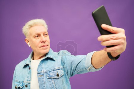 Téléchargez les photos : Homme âgé confiant avec les cheveux gris tenant smartphone dans la main tout en étirant le bras pendant la communication en ligne ou en prenant selfie - en image libre de droit
