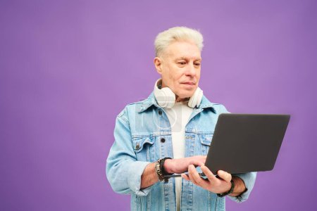 Téléchargez les photos : Homme d'affaires senior confiant avec des cheveux gris tapant sur le clavier de l'ordinateur portable tout en réseautant sur fond lilas en isolement - en image libre de droit