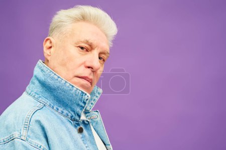 Téléchargez les photos : Homme âgé sérieux avec des cheveux blancs en jean bleu veste regardant la caméra tout en se tenant en isolement sur fond de lavande vive - en image libre de droit