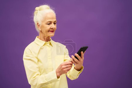 Photo for Senior woman in yellow shirt looking at smartphone screen while texting, communicating in video chat or watching online movie in isolation - Royalty Free Image