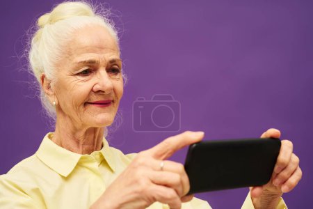 Téléchargez les photos : Femme âgée souriante aux cheveux blancs regardant l'écran du smartphone tout en regardant une vidéo ou un film en ligne curieux sur fond violet - en image libre de droit
