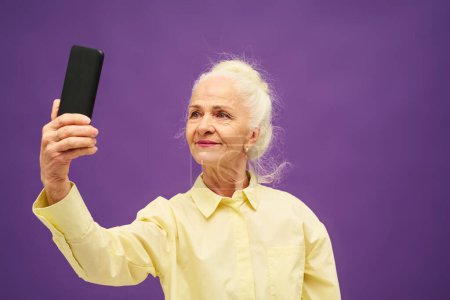 Téléchargez les photos : Femme âgée souriante en chemise jaune regardant l'écran du smartphone tout en prenant selfie ou en communiquant par chat vidéo sur fond violet - en image libre de droit