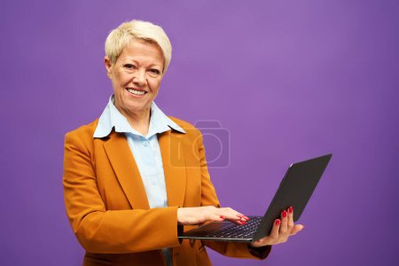Téléchargez les photos : Heureuse femme d'affaires mature en tenue de cérémonie regardant la caméra et regardant à travers les données en ligne dans l'ordinateur portable tout en se tenant dans l'isolement - en image libre de droit