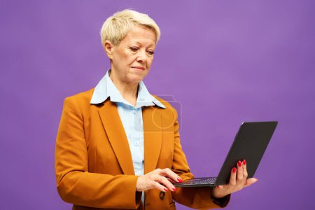 Téléchargez les photos : Femme d'affaires blonde senior avec ordinateur portable travaillant sur Internet ou recherchant des informations en ligne tout en regardant l'écran - en image libre de droit