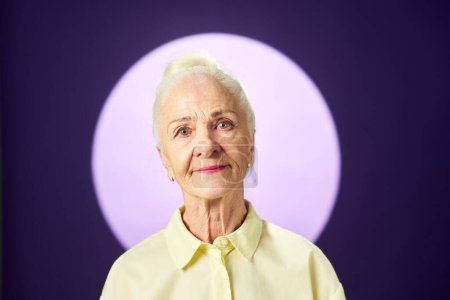 Téléchargez les photos : Femme âgée aux cheveux blancs regardant la caméra tout en se tenant contre la pleine lune sur fond de lavande et posant pendant la séance photo - en image libre de droit