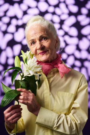 Foto de Mujer mayor sonriente con el pelo gris sosteniendo lirio blanco con hojas verdes y mirando a la cámara mientras posa sobre un fondo creativo - Imagen libre de derechos