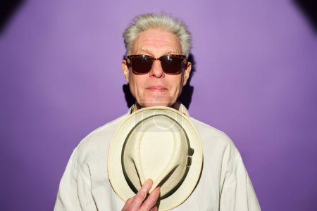 Téléchargez les photos : Homme mûr sérieux aux cheveux gris tenant un chapeau panama blanc par la poitrine tout en se tenant devant la caméra sur fond violet - en image libre de droit
