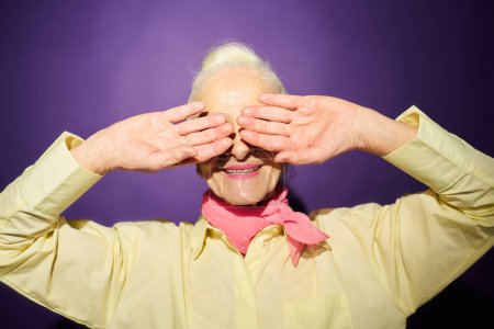Téléchargez les photos : Femme âgée souriante en chemise jaune couvrant les yeux par ses mains tout en se cachant de la lumière vive tout en se tenant debout sur fond de lavande - en image libre de droit