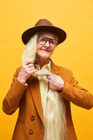 Téléchargez les photos : Femme âgée avec de très longs cheveux blancs debout devant la caméra, isolée sur fond jaune et posant pendant la séance photo - en image libre de droit