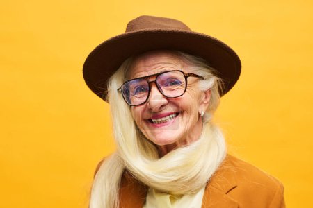 Téléchargez les photos : Portrait en gros plan d'une femme âgée joyeuse portant un chapeau et des lunettes en fedora marron avec ses longs poils blancs attachés autour du cou - en image libre de droit