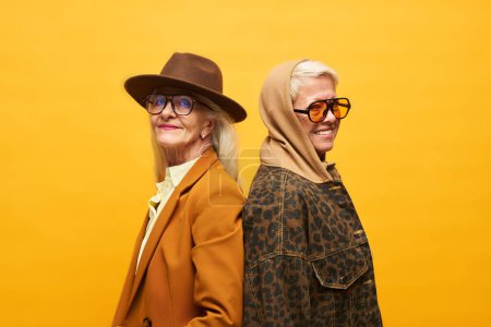 Foto de Dos mujeres mayores felices en ropa elegante posando frente a la cámara mientras están de pie sobre el fondo amarillo y tocando por la espalda - Imagen libre de derechos