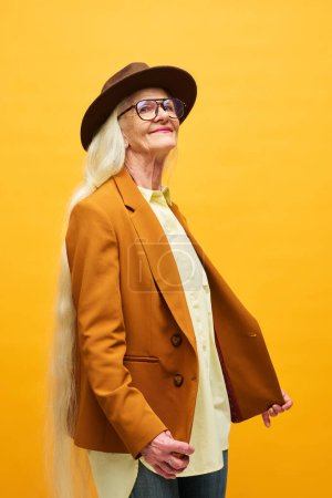 Foto de Mujer mayor feliz en gafas graduadas, chaqueta marrón y sombrero fedora mirando a la cámara mientras está de pie sobre el fondo amarillo durante la sesión de fotos - Imagen libre de derechos