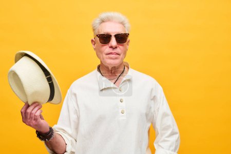 Téléchargez les photos : Homme sénior élégant en lunettes de soleil et chemise blanche tenant le chapeau panama dans la main droite tout en se tenant devant la caméra sur fond jaune - en image libre de droit