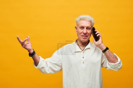 Téléchargez les photos : Heureux homme mature avec les cheveux gris parlant sur un téléphone portable et regardant la caméra tout en se tenant debout sur fond jaune dans l'isolement - en image libre de droit
