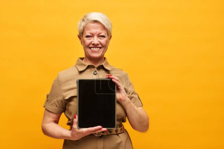 Photo for Cheerful mature woman with short blond hair holding tablet with blank screen while making presentation of project points in isolation - Royalty Free Image