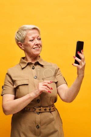 Photo for Stylish mature blond woman in dopamine dressing looking at smartphone screen while watching curious online video over yellow background - Royalty Free Image