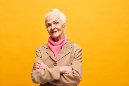 Foto de Mujer mayor en blazer beige y bufanda rosa cruzando los brazos por el pecho y mirando a la cámara mientras está de pie en aislamiento sobre el fondo amarillo - Imagen libre de derechos