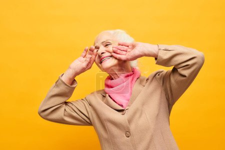 Téléchargez les photos : Joyeuse femme âgée en blazer beige et écharpe rose touchant le visage par les mains tout en riant devant la caméra sur fond jaune - en image libre de droit