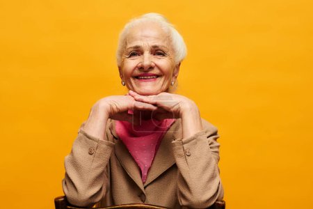 Téléchargez les photos : Femme âgée souriante en tenue décontractée intelligente assise devant la caméra en isolement tout en posant pendant la séance photo sur fond jaune - en image libre de droit