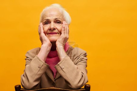 Foto de Elegante mujer mayor en blazer beige y bufanda rosa manteniendo las manos en las mejillas mientras se sienta en la silla en aislamiento y mirando a la cámara - Imagen libre de derechos
