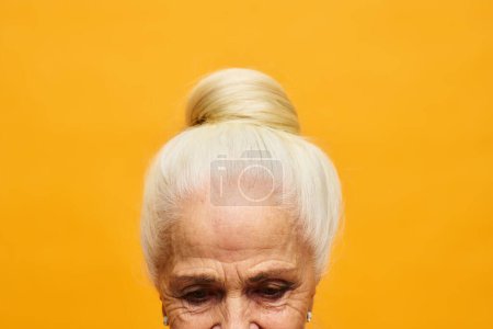 Téléchargez les photos : Plan recadré de la femme âgée aux cheveux blancs fait en chignon debout devant la caméra sur fond jaune et regardant vers le bas - en image libre de droit