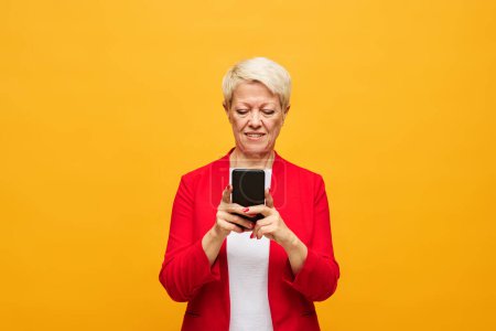 Photo for Aged blond woman with short hair looking at smartphone screen while texting or communicating in video chat over yellow background - Royalty Free Image