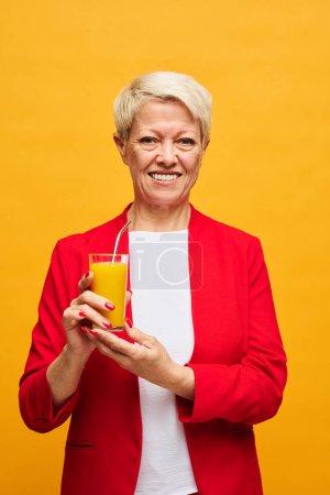 Foto de Mujer mayor feliz en chaqueta roja sosteniendo un vaso de jugo de naranja fresco y mirando a la cámara con sonrisa contra el fondo amarillo - Imagen libre de derechos