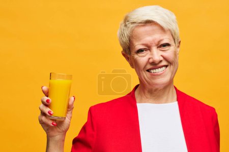 Téléchargez les photos : Femme blonde mature joyeuse avec sourire dents tenant un verre de jus d'orange et regardant la caméra tout en se tenant sur fond jaune - en image libre de droit
