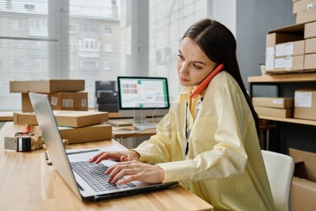 Téléchargez les photos : Bénévolat jeune femme brune occupée assise sur le lieu de travail devant un ordinateur portable et parlant sur un téléphone portable à des personnes en crise - en image libre de droit