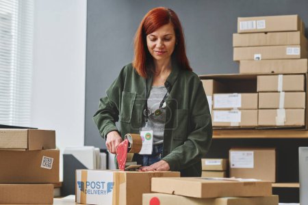 Photo for Young female volunteer sealing cardboard box with clothes with cellotape while standing by workplace with stack of packages and parcels - Royalty Free Image