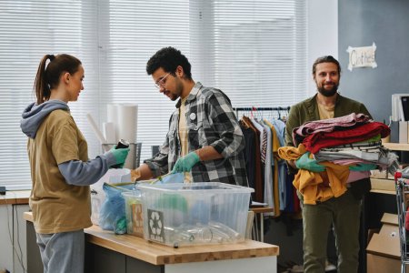 Photo for Two young volunteers sorting garbage while man carrying stack of second hand clothes while passing by behind them in volunteering office - Royalty Free Image