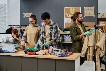 Photo for Group of young intercultural volunteers sorting second hand clothes on table and choosing casualwear on rack for people in need - Royalty Free Image