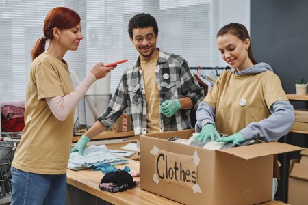 Photo for Young woman with smartphone taking photo of voluntters packing second hand clothes prepared for people in need in big cardboard box - Royalty Free Image