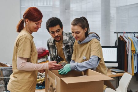 Photo for Happy woman showing two young intercultural volunteers photo of packing clothes in smartphone while standing by table with box - Royalty Free Image