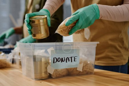 Foto de Primer plano de las manos enguantadas de voluntarios poniendo productos alimenticios en la caja de plástico, mientras que el embalaje de la donación para las personas necesitadas o refugiados - Imagen libre de derechos