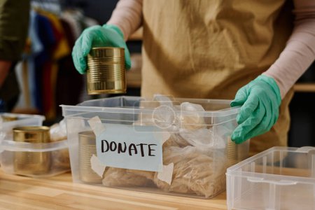 Photo for Gloved hands of young volunteer putting tinned food into plastic box while standing by table and preparing products for people in need - Royalty Free Image