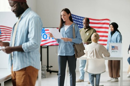 Foto de Mujer joven y feliz mirando a través de papeletas de votación mientras está de pie en la cola en el lugar de votación entre los ciudadanos interculturales de EE.UU. - Imagen libre de derechos